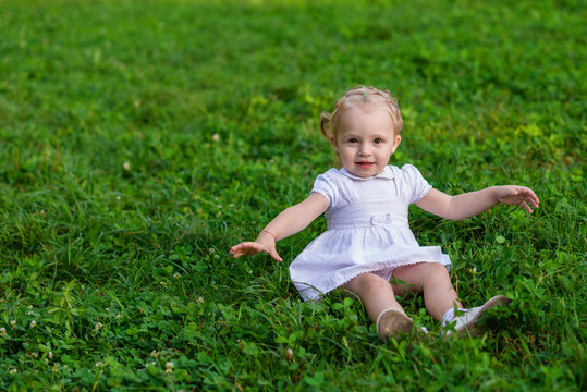 Baby girl in white dress sits and smiling on the fresh green grass