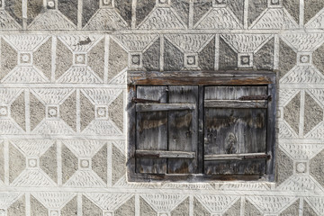 Eine Wand mit traditionellen Ornamenten und einem kleinen Fenster mit geschlossenen Holzläden an einem Haus im Bündnerland in der Schweiz.