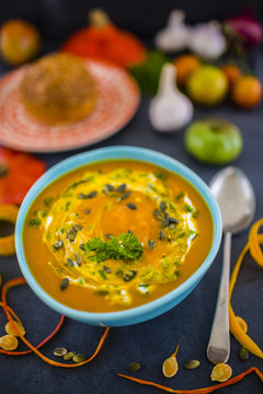 Cream of pumpkin soup with pumpkin seeds, white cream, vegetables in colourful bowl on black stone table.
