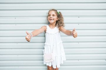 Little cute girl he put his hands forward and shows signs OK .  She looks away with a smile and dressed in a white summer dress .