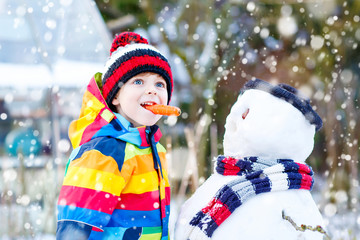 Funny kid boy in colorful clothes making a snowman, outdoors