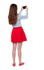 back view of standing young beautiful  woman  using a mobile phone. girl  watching. Rear view people collection.  backside view of person.  Isolated over white background. Long-haired brunette in red