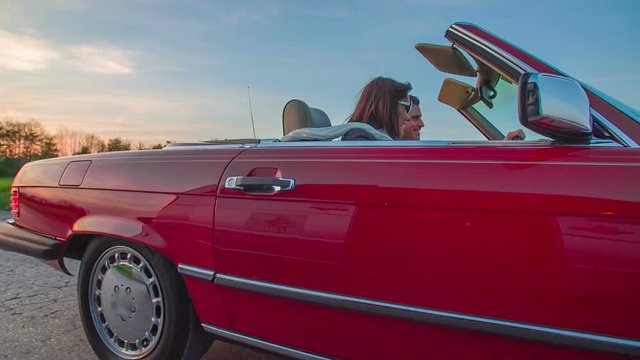 A couple is driving in a red car with open roof and they are having a wonderful time when travelling across the countryside. It is evening time. Close-up shot.
