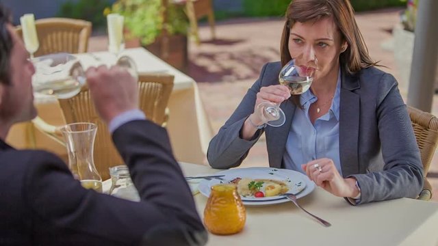 Two middle-aged people are enjoying their meal outside of a restaurant and they are smiling and drinking some white wine. Wide-angle shot.
