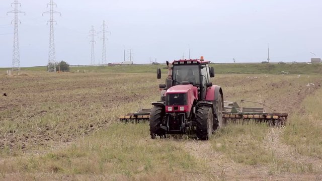 An agricultural tractor, plowing a field for sowing, moving to the camera. Birds are flying around