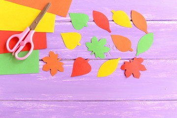 Leaves cut from colored paper, scissors, yellow, red, green and orange paper sheets on lilac wooden background. Children autumn art. Idea for a children pastime. Top view