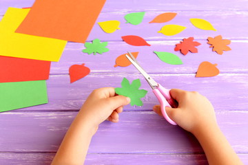 Child cuts out colored paper leaf. Child holds scissors and a green leaf in his hands. Colored paper set, colorful leaves on a wooden table. Autumn kids crafts idea