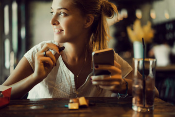 Young pretty woman using smartphone in cafe, drink coffee in cup, sweet breakfast, happy face, outdoor hipster portrait, fashion girl, table, sweet drink, tasty tea, aromatic coffee