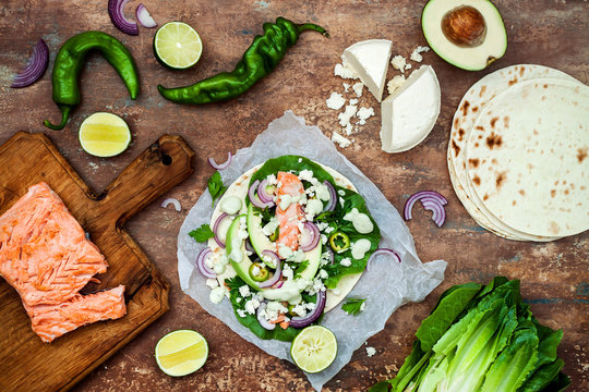 Preparing healthy lunch snacks. Fish tacos with grilled salmon, red onion, fresh salad leaves and avocado cilantro sauce on vintage stone background. Recipe for Cinco de Mayo party. Top view.