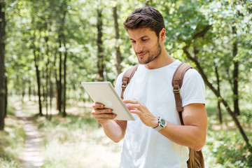 Fototapeta premium Smiling young man with backpack using tablet in forest
