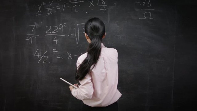  Teacher writing math formulas on blackboard & talking to her class