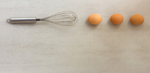 three chicken eggs and whisk on a steel gray background