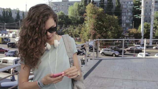 Young female brunette in sun glasses connect the headphones to the smart phone in the city, slow motion