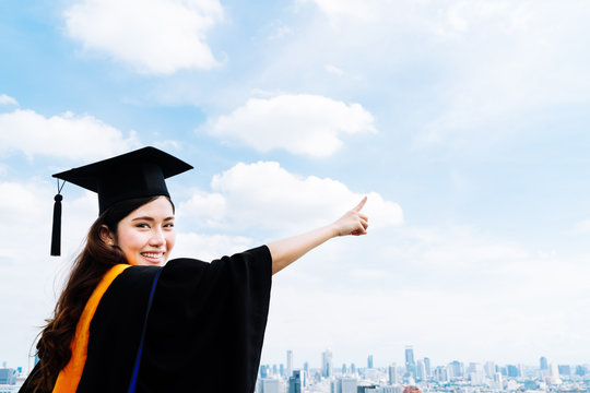 Beautiful asian university or college graduate student woman in graduation academic dress or gown, smiling and pointing at copy space on blue sky background, education or success concept