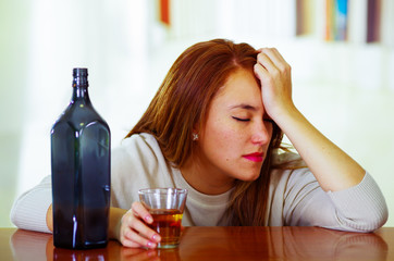 Attractive woman wearing white sweater sitting by bar counter lying over desk next to glass and bottle, drunk depressed facial expression, alcoholic concept