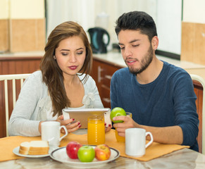 Young charming couple sitting by breakfast table looking at tablet screen, fruits, juice and coffee placed in front, hostel environment