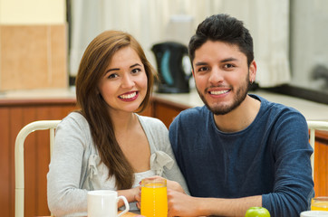 Young charming couple seated by breakfast table smiling to camera, fruits, juice and coffee placed in front, hostel environment