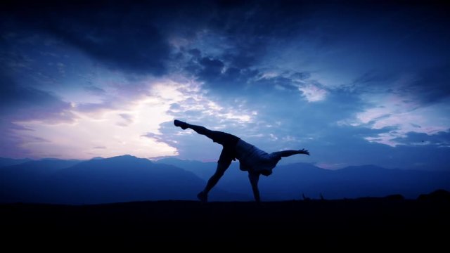 Silhouette of male acrobat doing a cartwheel on the top of a mountain. Slow motion