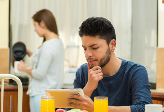 Young handsome man seated by breakfast table looking at tablet screen, girl standing behind pouring hot water, fruits, juice placed in front, hostel environment