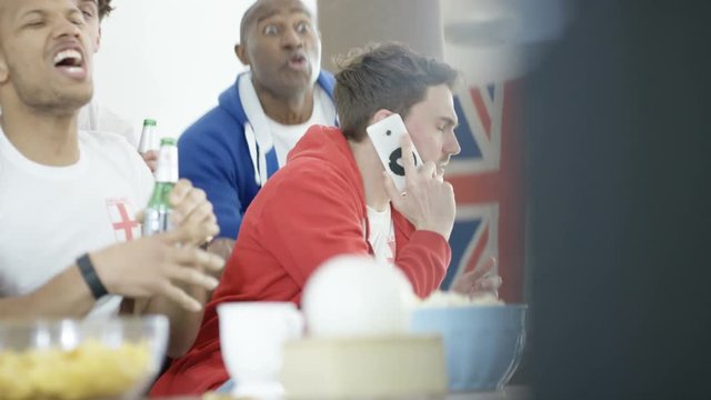  Man talking on mobile phone while rowdy friends watch sports game on TV