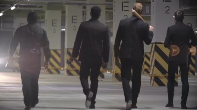 Slow motion rear view of four confident mafia men in black suits holding baseball bats and walking towards car with headlights on in empty parking lot.