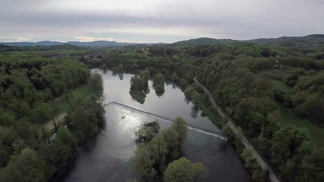 There is a river and there are many forests surrounding it. The day looks a bit rainy and not sunny. There are some mountains in the background, too. Wide-angle aerial shot.
