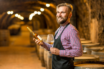 Portrait of a handsome sommelier in apron and checkered shirt with wine bottle standing in the old...