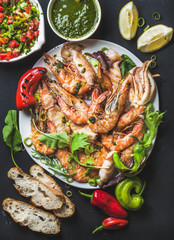 Plate of roasted tiger prawns and pieces of octopus with fresh leek, vegetable salad, peppers, lemon, bread and pesto sauce over black background, top view, vertical composition