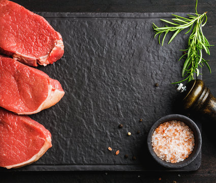 Raw beef Eye Round steaks with spices and rosemary on black slate stone board over dark wooden background, top view, copy space, horizontal composition