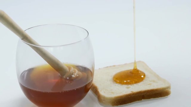 Some honey is in a cup together with a wooden honey stick. A piece of bread is standing on a kitchen counter and some honey is pouring down on it. Close-up shot.
