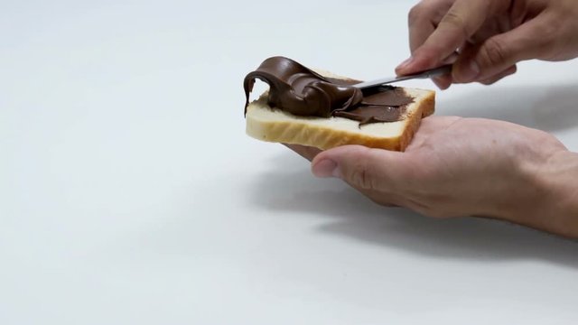 A man is holding a piece of toast in his hand and he is trying to spread out Nutella on it. Close-up shot.
