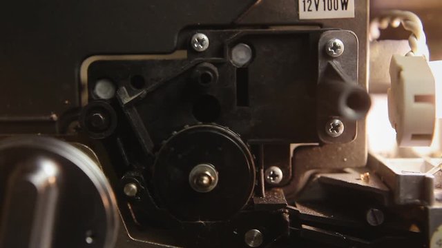 Inside a super 8mm film projector: a spinning wheel, the lamp, the on-off button. A hand turns off the machine. Macro detail angled shot.
