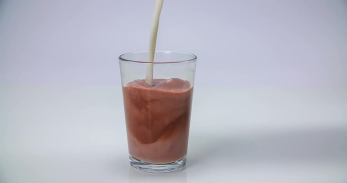 A glass with some chocolate drink is standing on a table and someone is adding some more milk into it. Close-up shot.
