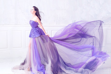Beautiful brunette woman wearing long purple dress posing in empty light interior