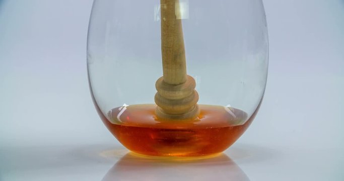 A glass with little honey in it is standing on a kitchen counter. Suddenly, a wooden honey stick falls into it. Close-up shot.
