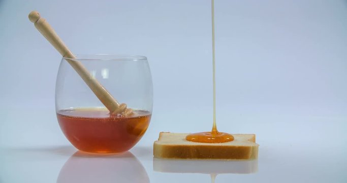 A piece of toast and a glass with honey are standing on a table. Someone starts pouring some honey on the top of this toast. Wide-angle shot.
