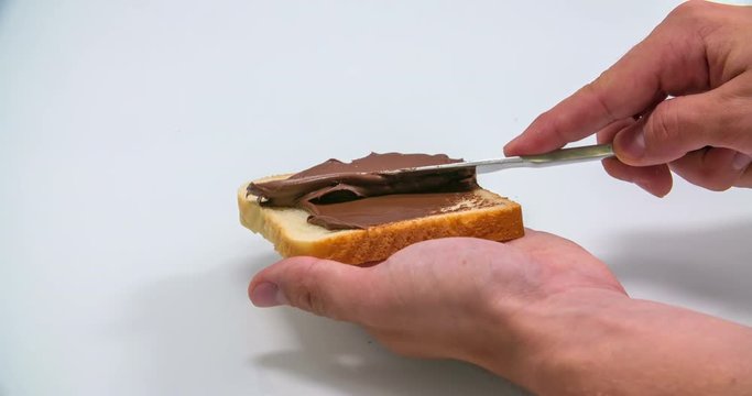 A man is holding a piece of toast in his hand and he is slowly spreading out some nutella on it. Close-up shot.
