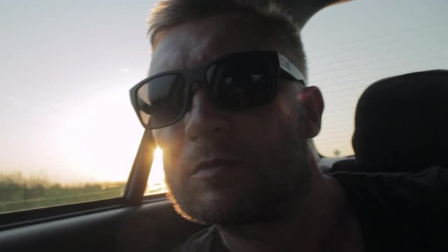 Young casual man with beard portrait sitting on the backseat of car. Urban male hipster in his 30s. Sunset
