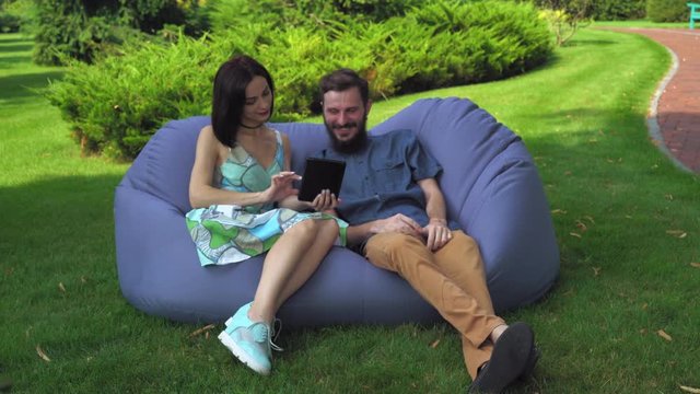 A young couple in bright clothes sits in a chair ottoman in the park on a summer day and browsing photos on the tablet.