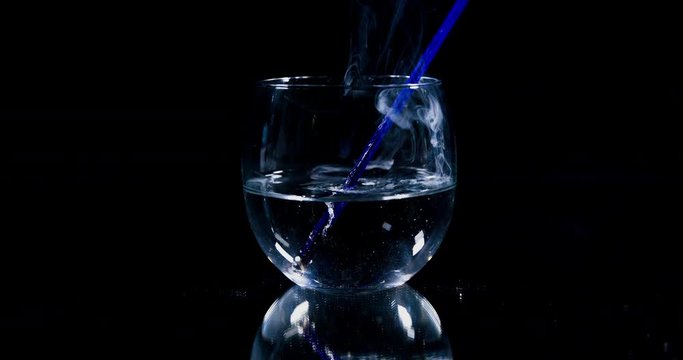 A glass of water is standing on the table in the kitchen. A blue straw and a burning match fall into it. We can see a smoke when a match dies out.
