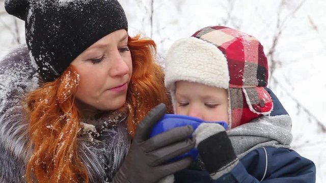 Close up video of young happy mother and little son drinking hot tea from one plastic big cup outdoors. Head and shoulders portrait of smiling and laughing family on winter picnic on holiday.