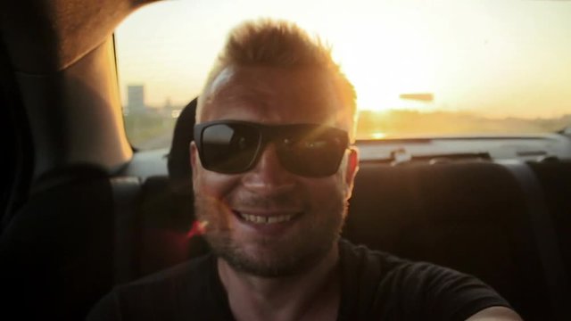 Young casual man with beard portrait sitting on the backseat of car. Urban male hipster in his 30s. Handsome young beared man smiling happy outside looking at camera. Sunset