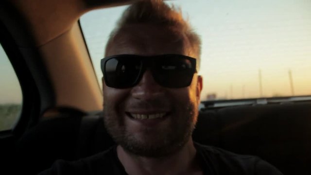 Young casual man with beard portrait sitting on the backseat of car. Urban male hipster in his 30s. Handsome young beared man smiling happy outside looking at camera. Sunset