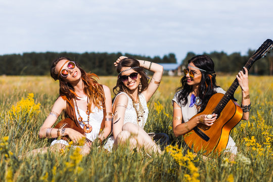 Three cute hippie girl sitting on grass outdoors, best friends having fun and laughing, playing the guitar, sunglasses, feathers in their hair, bracelets, flash tattoo, indie, Bohemia, boho style
