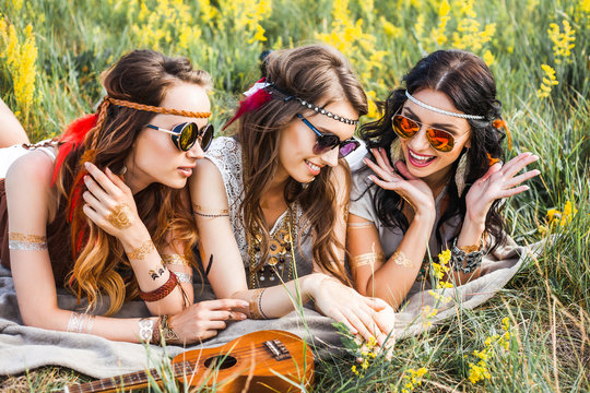 Three cute hippie girl lying on the grass among the wildflowers, best friends having fun and laughing, ukulele, sunglasses, feathers in their hair, bracelets, flash tattoo, indie, Bohemia, boho style