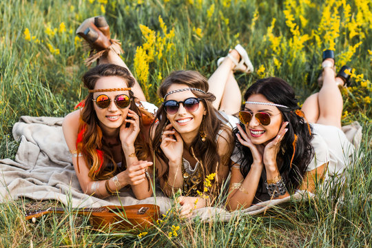 Three cute hippie girl lying on the grass among the wildflowers, best friends having fun and laughing, accessories sunglasses, feathers in their hair, bracelets, flash tattoo, indie Bohemia boho style
