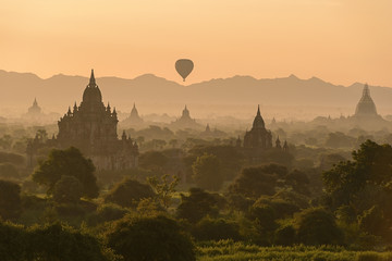 A colorful hot air balloon floats over a lush green forest, with  temples peeking through the trees.  In the distance, there are  mountain ranges.