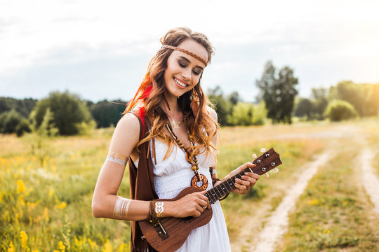 Pretty amazing free red-haired hippie girl playing a ukulele outdoors, feathers and braids in her hair, white dress, leather and gold accessories, flash tattoo, indie, Bohemia, boho style, nice smile