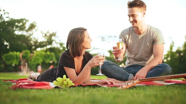 Young beautiful couple smiling, drinking champagne, resting on picnic in park. Slow motion.