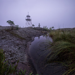 Obraz premium Misty landscape with an old lighthouse covered by fog at Kurkiniemi cape at the north of Ladoga lake. On the foreground a bush of reed with many rain drops on its leafs grow on the edge of small pond.